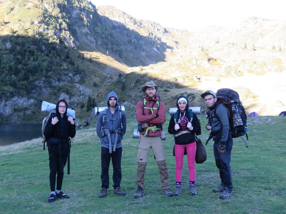 Cinq membres du club, sac à dos et tapis de sol, devant un lac de montagne lors d'un week-end